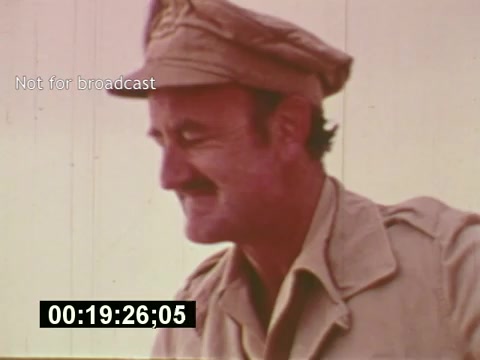 The still from 'Sixty Second Spot' captures a moment in the production of a 7-Up commercial, showcasing the behind-the-scenes process from script development to filming. The image features a man in a military-style cap and khaki attire, likely a crew member, smiling as he interacts on set. The frame includes a timecode and a watermark indicating it is not for broadcast, emphasizing its documentary nature. This segment serves to illustrate the creative and logistical efforts involved in commercial filmmaking during the mid-1970s.