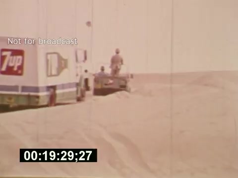 The still from 'Sixty Second Spot' depicts a behind-the-scenes moment in the creation of a 7-Up commercial from the mid-1970s. It features a truck adorned with the 7-Up logo parked in a sandy desert landscape. Two individuals, one on foot and one on a vehicle, appear to be involved in the filming process. The image captures the effort behind commercial production, reflecting the era's aesthetic and the outdoor setting used for the shoot. The timecode at the bottom suggests it's part of a longer sequence, showcasing the stages of development from concept to execution.