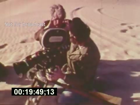 The still depicts a behind-the-scenes moment from 'Sixty Second Spot,' showcasing the filming process of a 7-Up commercial from the mid-1970s. Two crew members are seen working with a 16mm camera in a sandy location, capturing the essence of the commercial's creation. The timestamp at the bottom indicates a specific moment in the film, while the 'Not for broadcast' label suggests this content is for reference purposes only. Overall, the image provides a glimpse into the filmmaking process, including script development, casting, and technical execution.
