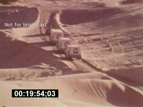The 16mm still from 'Sixty Second Spot' depicts a scene from the making of a 7-Up commercial in the mid-1970s. It features several trucks traversing a desert landscape, emphasizing the logistical aspects of commercial production. The timestamp at the bottom suggests a specific moment in the film, while the 'Not for broadcast' label indicates it’s a behind-the-scenes look, capturing the creative process behind the final advertisement.