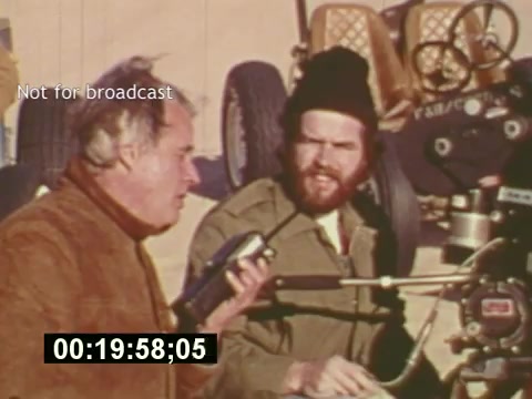 The still from 'Sixty Second Spot' captures a moment during the production of a 7-Up commercial in the mid-1970s. It depicts two men engaged in conversation, likely discussing the filming process. One is using a microphone, possibly providing directions or commentary, while the other listens attentively. The background suggests an outdoor filming set, complete with equipment. The scene highlights the collaborative effort involved in creating a television advertisement.