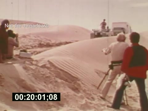 The still from 'Sixty Second Spot' captures a behind-the-scenes moment in the creation of a 7-Up commercial. Filmed in a desert setting, it shows crew members actively engaged in filming. You can see a person in a vehicle, possibly a director or camera operator, guiding the shoot, while others set up equipment and capture footage. The image conveys the collaborative effort and dynamic environment of commercial production during the mid-1970s. The timestamp indicates an ongoing scene, showcasing the process of bringing a script to life.