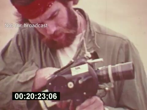 The still from 'Sixty Second Spot' features a filmmaker in mid-1970s attire, concentrating on a 16mm camera. His rugged look, complete with a beard and headband, reflects the creative, hands-on spirit of the era. The backdrop is simple, emphasizing the subject's focus on the filmmaking process. This moment encapsulates the behind-the-scenes work involved in creating a 7-Up commercial, showcasing the journey from script to screen. The timestamp suggests it's part of a documentary-style exploration of television advertising.