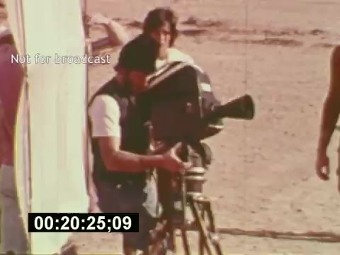 The still from 'Sixty Second Spot' captures a behind-the-scenes moment in the making of a 7-Up commercial from the mid-1970s. It showcases a camera operator setting up a 16mm film camera on a tripod, with a crew member in the background. The sandy environment suggests an outdoor shoot, emphasizing the film's focus on the commercial's production process, from script to filming. The timestamp and 'Not for broadcast' label indicate this footage is meant for archival purposes rather than public airing.