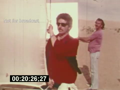 This still from 'Sixty Second Spot,' a film detailing the creation of a 7-Up commercial, depicts two men on location. One man, dressed in a red shirt and sunglasses, is engaged in adjusting a lighting setup, while another man in a pink shirt assists in the background. The sandy environment suggests an outdoor shoot. The film highlights the process of bringing a commercial to life, from scripting to casting.