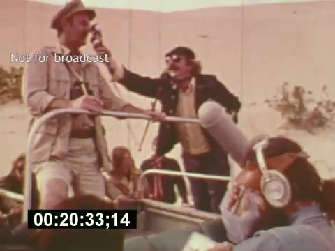 The still from 'Sixty Second Spot' captures a behind-the-scenes moment in the creation of a 7-Up commercial. It features a group of filmmakers in a desert setting, with a man in a hat directing while another person holds a microphone, suggesting a lively production atmosphere. Crew members can be seen seated, illustrating the collaborative effort involved in the filming process. This snapshot provides a glimpse into the behind-the-scenes dynamics of 1970s commercial filmmaking.