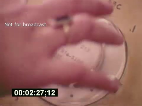 The still from 'Sixty Second Spot' features a hand approaching a circular object, likely a film reel or projector plate, with a timestamp indicating the scene time. The caption 'Not for broadcast' suggests this is a behind-the-scenes moment, emphasizing the documentary nature of the film. The context revolves around the production process of a 7-Up commercial from the mid-1970s, showcasing elements like script development, casting, and filming.