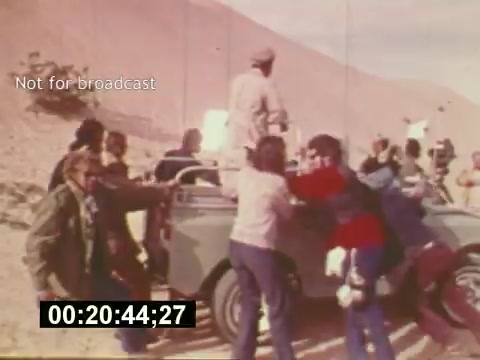 The still from 'Sixty Second Spot' captures a behind-the-scenes moment from the filming of a 7-Up commercial in the mid-1970s. The scene depicts a bustling crew working around a vintage vehicle in a desert-like setting. Several people are visible, some engaged in directing and others assisting with equipment, illustrating the collaborative effort that goes into creating a commercial. The timestamp indicates it’s a pivotal moment, reflecting the dynamic and energetic atmosphere typical of film productions. The label 'Not for broadcast' suggests this footage is intended for internal use, providing a glimpse into the filmmaking process rather than the final product.