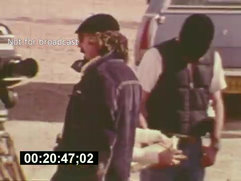 The still from 'Sixty Second Spot' captures a behind-the-scenes moment in the making of a 7-Up commercial from the mid-1970s. It shows a director or crew member engaged with the camera setup, while another individual adjusts equipment or props. The scene highlights the collaborative and dynamic atmosphere of film production during that era, showcasing a mix of creativity and technical expertise essential in crafting commercials. The 'Not for broadcast' watermark indicates this content is exclusive to the film's production context.