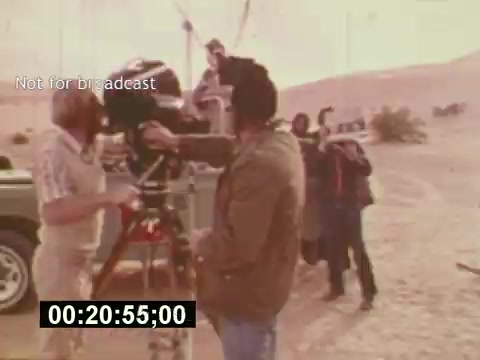 The still from 'Sixty Second Spot' captures a behind-the-scenes moment in the creation of a 7-Up commercial from the mid-1970s. It features a director or cinematographer adjusting a camera on a tripod, while crew members are set up in a desert-like location. The atmosphere appears casual yet focused, highlighting the collaborative effort involved in the filmmaking process. The timestamp and 'Not for broadcast' label indicate it's an outtake or production footage rather than a finished product.