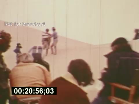 The still from 'Sixty Second Spot' captures a scene from the making of a 7-Up commercial in the mid-1970s. It features a film set with individuals involved in the production process, including crew members and cast. The background suggests a sand dune location, while the foreground shows people seated, possibly directors or crew, watching the filming unfold. The image reflects the collaborative effort of filmmaking, showcasing behind-the-scenes elements typical of commercial production.