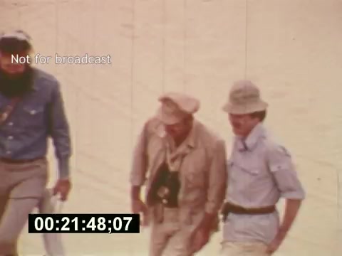 The 16mm still from 'Sixty Second Spot' captures a moment during the making of a 7-Up commercial in the mid-1970s. It features three men walking together in a sandy environment, dressed in casual attire suitable for filming. The scene reflects the behind-the-scenes process of commercial creation, emphasizing the collaborative efforts involved in script development, casting, and filming. The timestamp at the bottom indicates its position within the film, while the 'Not for broadcast' label suggests it is a production still rather than a public release.