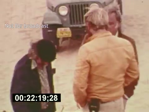 The 16mm still from 'Sixty Second Spot' captures a moment in the creation of a 7-Up commercial in the mid-1970s. It features a group of individuals, likely involved in the production process, gathered in a sandy outdoor location. One person wears a yellow jacket, indicating a leadership or key role, while others appear to be engaged in discussion or reviewing materials. A vintage Jeep is parked behind them, adding to the retro feel of the scene. The still emphasizes the behind-the-scenes aspect of commercial filmmaking, showcasing the collaborative nature of the creative process.