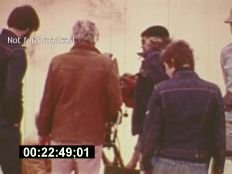 The still from 'Sixty Second Spot' captures a behind-the-scenes moment during the creation of a 7-Up commercial from the mid-1970s. It features a small group of individuals, likely including crew members and cast, gathered around filming equipment. The casual attire, including denim jackets, reflects the era's style. The image conveys a sense of teamwork and the creative process involved in commercial production, highlighting the transition from script to screen. A timestamp indicating the scene's position within the film is visible at the bottom.