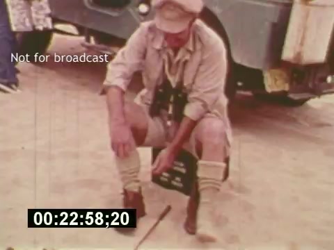 The 16mm still from 'Sixty Second Spot' captures a behind-the-scenes moment in the creation of a 7-Up commercial from the mid-1970s. It features a crew member dressed in period attire, squatting while holding a clapboard, signaling the start of a take. The sandy setting, combined with a vintage vehicle in the background, sets the scene for the filming process. The timecode displayed suggests it's part of a documentary style, highlighting the meticulous nature of commercial production.