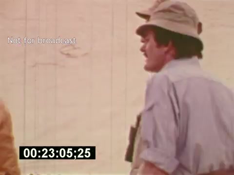 The still from 'Sixty Second Spot' captures a behind-the-scenes moment during the creation of a 7-Up commercial from the mid-1970s. It features a man in a hat, possibly a director or crew member, standing against a sandy backdrop. The image seems to illustrate the production process, highlighting the effort and attention to detail involved in commercial filmmaking. The timecode indicates it’s part of a larger sequence, emphasizing its documentary style.