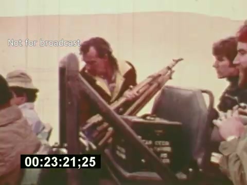 The still from 'Sixty Second Spot' captures a behind-the-scenes moment in the making of a 7-Up commercial from the mid-1970s. It features a crew of filmmakers, with one member focusing on a camera setup while others look on. The environment hints at an outdoor shoot, likely in a remote location. This snapshot showcases the collaborative effort and technical aspects involved in commercial filmmaking during that era, emphasizing the production process from script to screen.