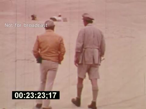 The still captures two men walking in a sandy, outdoor setting, possibly a desert. One man is dressed in a light-colored jacket, while the other wears a hat and shorts, suggesting a casual, warm-weather vibe. The scene appears to be part of the behind-the-scenes process of creating a 7-Up commercial in the mid-1970s. The timecode at the bottom indicates it's a film still from 'Sixty Second Spot,' focusing on the development and production of the advertisement.