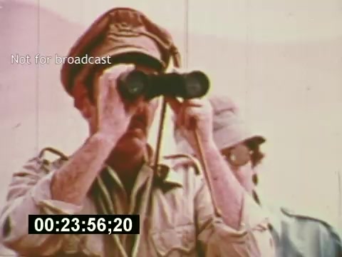 The still from 'Sixty Second Spot' features an outdoor scene where a man in military attire, wearing a cap, is using binoculars. He appears focused on something in the distance, while another person, slightly behind him, is also visible. The image captures a moment that aligns with the film's exploration of the behind-the-scenes process in creating a 7-Up commercial, showing the planning and attention to detail involved. The timecode indicates it is a specific moment in the film but is not for broadcast.