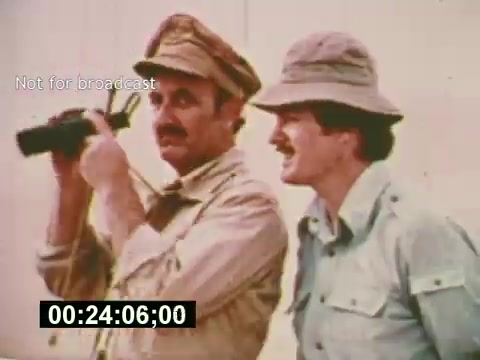 The still from 'Sixty Second Spot' features two men in outdoor attire, one using binoculars and the other standing beside him. The scene captures a moment during the production of a 7-Up commercial, highlighting the behind-the-scenes process of casting and filming in the mid-1970s. The men appear engaged and focused on their task, reflecting the creative efforts involved in commercial filmmaking.