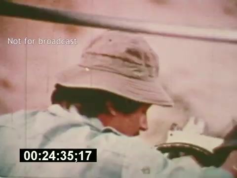 The 16mm still from 'Sixty Second Spot' captures a moment during the creation of a 7-Up commercial from the mid-1970s. It shows a crew member, wearing a hat, focused on the filming process. The image reflects the behind-the-scenes efforts in casting and production, highlighting the meticulous work involved in commercial filmmaking. The timecode displayed emphasizes this is a segment from the film, which contrasts the creative process with the final product that is shown at the film's conclusion.