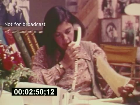 The still captures a scene from the film 'Sixty Second Spot,' focusing on the behind-the-scenes process of creating a 7-Up commercial. It features a woman engaged in a phone conversation, likely discussing details related to the commercial's production. The setting appears to be a creative or office space, adorned with photographs, emphasizing the collaborative environment of advertising in the mid-1970s. The timecode suggests this moment falls early in the film, outlining the initial stages of production.