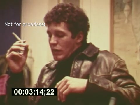 The 16mm still captures a scene from 'Sixty Second Spot,' a film that delves into the creation of a 7-Up commercial from the mid-1970s. The image features a young man in a leather jacket, smoking a cigarette, presumably during a discussion about the commercial's development. The setting has an informal, behind-the-scenes vibe, and the timestamp indicates it’s a moment caught early in the production process, reflecting the creative atmosphere of the time.