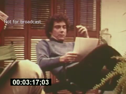 The still from 'Sixty Second Spot' captures a moment in the creation of a 7-Up commercial from the mid-1970s. It features a young man sitting in a relaxed manner, reviewing a script. The setting includes wooden shutters and greenery, suggesting a casual working environment. The time code indicates a specific point in the film, highlighting a behind-the-scenes look at the casting and filming process. This frame encapsulates the creative effort involved in advertising during that era.