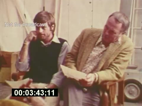 The still captures a behind-the-scenes moment from the film 'Sixty Second Spot,' focusing on the making of a 7-Up commercial in the mid-1970s. It features two men: one appears contemplative with a thoughtful expression, while the other, dressed in a tweed jacket, closely examines a script. The setting suggests a relaxed, yet professional atmosphere typical of a film set, with elements indicative of the production process visible in the background. The timestamp indicates this is a specific moment in the film, likely emphasizing discussion or decision-making during the creative process.