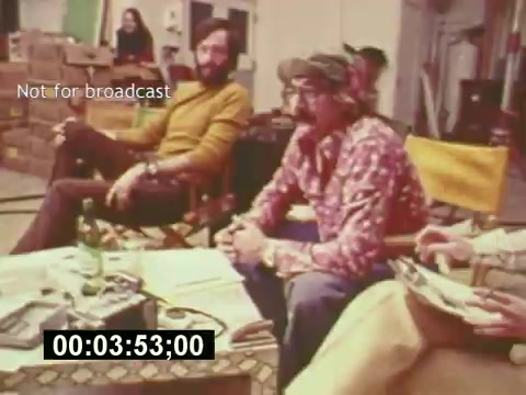The still from 'Sixty Second Spot' captures a behind-the-scenes moment in the creation of a 7-Up commercial from the mid-1970s. It shows a group of individuals, likely cast and crew members, seated around a table filled with production materials. The environment is casual, indicative of a creative brainstorming session. The prominent 'Not for broadcast' label suggests this is a raw, unedited snippet, providing an insider's look at the filmmaking process, emphasizing the collaborative effort involved in advertising creation. A timer at the bottom indicates the duration of the clip.