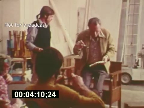 This still from 'Sixty Second Spot' captures a behind-the-scenes moment in the creation of a 7-Up commercial from the mid-1970s. The scene shows a director or producer seated, discussing with a crew member, while another individual appears to be engaged in the discussion. The casual yet focused atmosphere highlights the collaborative nature of commercial filmmaking during that era, with various elements of the production process visibly unfolding. The timestamp suggests it’s a key moment in the documentary, emphasizing the behind-the-scenes efforts involved in crafting advertising content.