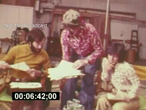 The still captures a behind-the-scenes moment from the film 'Sixty Second Spot,' showcasing the collaborative process of creating a 7-Up commercial in the mid-1970s. Three individuals—two men and one woman—are seated and engaged in discussion, surrounded by paperwork and notes. The man in a colorful, patterned shirt is actively pointing at the script while the others listen attentively, suggesting a brainstorming or planning session. The setting appears informal, likely in a studio or production area, reflecting the creative energy and teamwork involved in commercial filmmaking. The timestamp indicates this scene occurs 6 minutes and 42 seconds into the film.