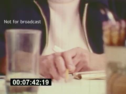 The still from 'Sixty Second Spot' captures a moment in the creation of a 7-Up commercial from the mid-1970s. It features a close-up of a hand holding a pencil, poised over a notepad, suggesting brainstorming or scriptwriting. In the background, there's a glass and some objects hinting at a creative workspace. The frame is marked 'Not for broadcast,' indicating its behind-the-scenes nature. This snapshot reflects the collaborative process of commercial filmmaking, emphasizing the artistic and planning phases before the final product is showcased at the film's end.