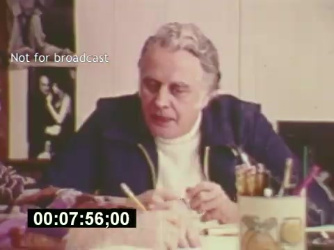 The still from 'Sixty Second Spot' features a man in a casual outfit, seated at a table cluttered with papers and writing materials. He appears to be involved in a creative discussion, likely about the production of a 7-Up commercial. In the background, there are posters and images that reflect the marketing context of the mid-1970s. The scene captures the behind-the-scenes process of script development, casting, and filming, providing a glimpse into the collaborative nature of commercial creation. A timer at the bottom suggests a specific moment in the film.