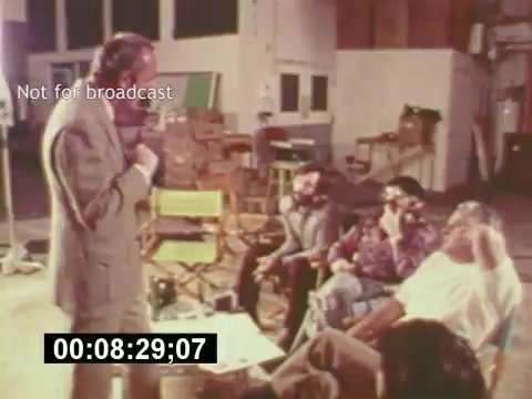 The still from 'Sixty Second Spot' captures a behind-the-scenes moment in the creation of a 7-Up commercial from the mid-1970s. In a studio setting, a director or producer, dressed in a suit, stands and addresses a small audience of crew members seated casually. The atmosphere is relaxed yet focused, indicative of the collaborative effort involved in filmmaking. This provides insight into the casting, scripting, and production process, emphasizing the creative dynamics of commercial filmmaking. The timestamp and 'Not for broadcast' label signify its intended use as educational or archival material.