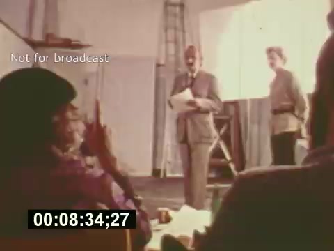 The still from 'Sixty Second Spot' showcases a behind-the-scenes moment during the creation of a 7-Up commercial from the mid-1970s. A man in a suit holds a script, addressing a small group seated in the foreground, while a woman stands beside him. The setting appears to be a studio, with visible production equipment in the background. This captures the collaborative atmosphere of casting and filming, highlighting the creative process involved in commercial production.