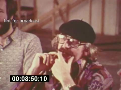 The still from 'Sixty Second Spot' captures a moment during the production of a 7-Up commercial, showcasing behind-the-scenes interactions in the mid 1970s. It features a man with glasses and a beret, engaged in thought while seated next to another individual, suggesting a creative brainstorming or discussion process. The image conveys a sense of nostalgia for the era of commercial filmmaking, emphasizing the collaborative effort involved in bringing the advertisement to life. A timecode at the bottom indicates the specific moment in the film. The overlay 'Not for broadcast' highlights that this content is intended for reference rather than public viewing.