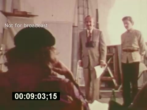 The still from 'Sixty Second Spot' captures a moment during the production of a 7-Up commercial in the mid-1970s. It features a scene where two men, dressed in formal and casual attire, appear to be engaging in a discussion or presentation, while a third person is partially visible, likely observing. The setting suggests a creative environment, possibly a casting or brainstorming session, illuminated by natural light. The text 'Not for broadcast' indicates its use for behind-the-scenes purposes, reflecting on the process of commercial filmmaking.