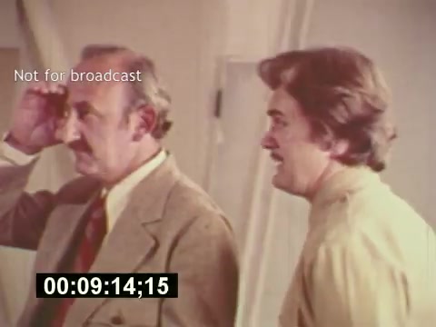 The still from 'Sixty Second Spot' features two men engaged in conversation, likely discussing the creation of a 7-Up commercial. One man, dressed in a suit, appears contemplative, while the other, in a lighter shirt, seems animated and engaged. The shot emphasizes the behind-the-scenes process of commercial filmmaking in the mid-1970s, highlighting elements such as casting and script development. The timestamp indicates it's a specific moment in the film, which captures the creative dynamics of advertising during that era.