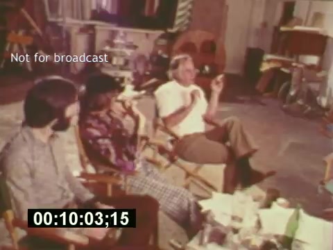 This 16mm still from 'Sixty Second Spot' captures a moment during the creation of a 7-Up commercial in the mid-1970s. It features a group of three individuals seated in director's chairs, engaged in discussion on a film set. The atmosphere is informal, with various items scattered on the table, suggesting a creative brainstorming session. The timestamp and 'Not for broadcast' watermark indicate its purpose as behind-the-scenes footage rather than a final product. This snapshot reflects the collaborative process of commercial filmmaking during that era.