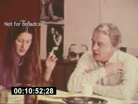 The still from 'Sixty Second Spot' captures a moment in the creation of a 7-Up commercial from the mid-1970s. It features a woman with long hair, holding a cigarette, and a man in a patterned shirt, both engaged in discussion. The setting suggests a creative environment, with notes and scripts visible, highlighting the collaborative process of casting and filming for the ad. This behind-the-scenes glimpse illustrates the intricate development involved in producing a TV commercial.