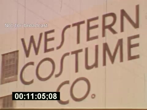 The still from 'Sixty Second Spot' features a prominent building facade with the sign 'WESTERN COSTUME CO.' It reflects the mid-1970s production process of a 7-Up commercial, highlighting the behind-the-scenes elements such as script development, casting, and filming. The visual emphasizes the importance of costume design in the creation of advertisements during that era.