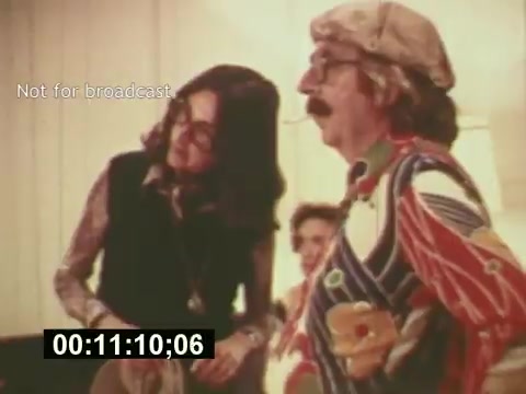 The still captures a scene from 'Sixty Second Spot,' showcasing the behind-the-scenes creation of a 7-Up commercial from the mid-1970s. It features two individuals engaging in conversation, with the man on the right wearing a colorful patterned shirt and a hat, while the woman on the left appears to be observing intently. The atmosphere reflects a casual, creative setting typical of film production during that era. A timecode at the bottom indicates the specific moment in the film, suggesting it's a shot from the editing process.