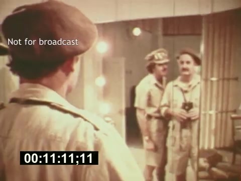 The still from 'Sixty Second Spot' features two characters in a dressing room, preparing for a 7-Up commercial. One actor stands in front of a mirror, reflecting a moment of anticipation, while the other appears to engage with him, likely offering guidance or support. The vintage aesthetic emphasizes the mid-1970s production vibe, capturing the behind-the-scenes process of commercial filmmaking. The image conveys a mix of excitement and the collaborative nature of creative work in advertising.