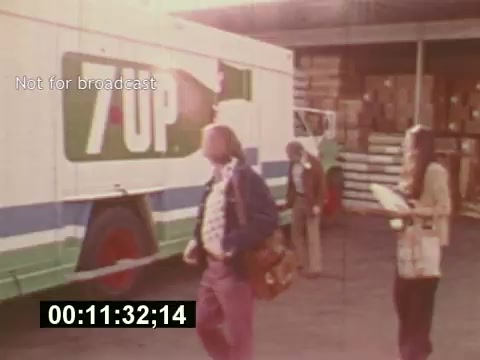The still from 'Sixty Second Spot' depicts a behind-the-scenes moment in the creation of a 7-Up commercial from the mid-1970s. It features a delivery truck marked with the 7-Up logo parked in a warehouse setting. Individuals, including a man with a bag and a woman holding a script, are seen discussing and preparing for the shoot. The scene captures the essence of the commercial's development process, highlighting elements of casting and filming. A timecode at the bottom indicates the specific segment of footage.