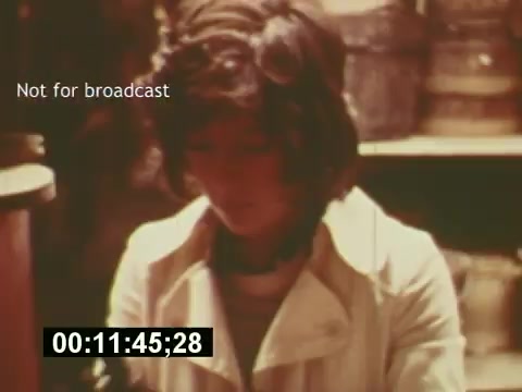 The still from 'Sixty Second Spot' captures a moment in the process of creating a 7-Up commercial. It features a young woman in a light-colored coat, appearing focused as she works. The background suggests a cluttered space, likely related to the filming. The timestamp and 'Not for broadcast' note indicate its production use while hinting at the behind-the-scenes nature of the project, which details the commercial's creation from script to screen.