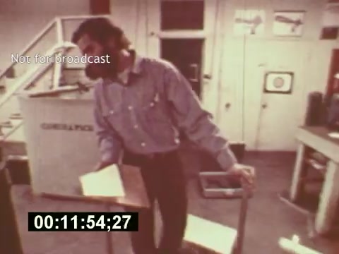 The still from the film 'Sixty Second Spot' depicts a behind-the-scenes moment in the creation of a 7-Up commercial from the mid-1970s. A man in a casual shirt is seen interacting with props or scripts in a production environment, surrounded by equipment typical of the era. The scene emphasizes the collaborative and detailed process of advertising production, showcasing the planning and creativity involved in bringing a commercial to life. The timestamp suggests it's a segment showcasing the development phase leading up to the final advertisement.