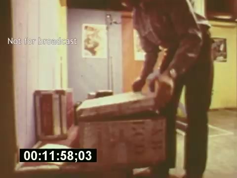 The still from 'Sixty Second Spot' likely depicts a behind-the-scenes moment in the creation of a 7-Up commercial from the mid-1970s. It shows a person handling a box, possibly containing props or materials for the shoot. The setting appears informal, with posters visible on the walls, highlighting the creative environment. The timestamp indicates the specific moment in the film, suggesting a focus on the production process. This scene emphasizes the collaborative effort involved in developing a commercial, from script to casting and filming.