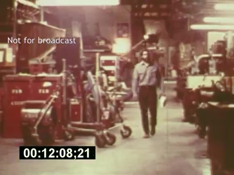 The still from 'Sixty Second Spot' depicts a behind-the-scenes moment in a film studio, likely during the mid-1970s. The scene features a man walking through a cluttered workspace filled with equipment and production tools, emphasizing the industrial environment of commercial filmmaking. The text 'Not for broadcast' suggests that this footage is intended for internal use, showcasing the creative process behind a 7-Up commercial. The timecode at the bottom indicates the timing within the film, reinforcing its documentary style.