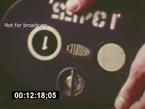 The still captures a moment from 'Sixty Second Spot,' focusing on the behind-the-scenes process of creating a 7-Up commercial in the mid-1970s. It showcases a close-up of equipment, highlighting a hand operating a reel marked with '7-Up.' The scene emphasizes the technical aspects of filming, set against the backdrop of the commercial's development, from scripting to casting, eventually leading to the screening of the finished ad. The timestamp indicates this is part of the documentary's exploration of television advertising production.